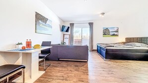 Ein modernes Zimmer mit Schreibtisch, Bett und Schlafsofa. Zwei Fenster mit Vorhängen, ein Flachbildfernseher und ein gerahmtes Bild an der Wand.