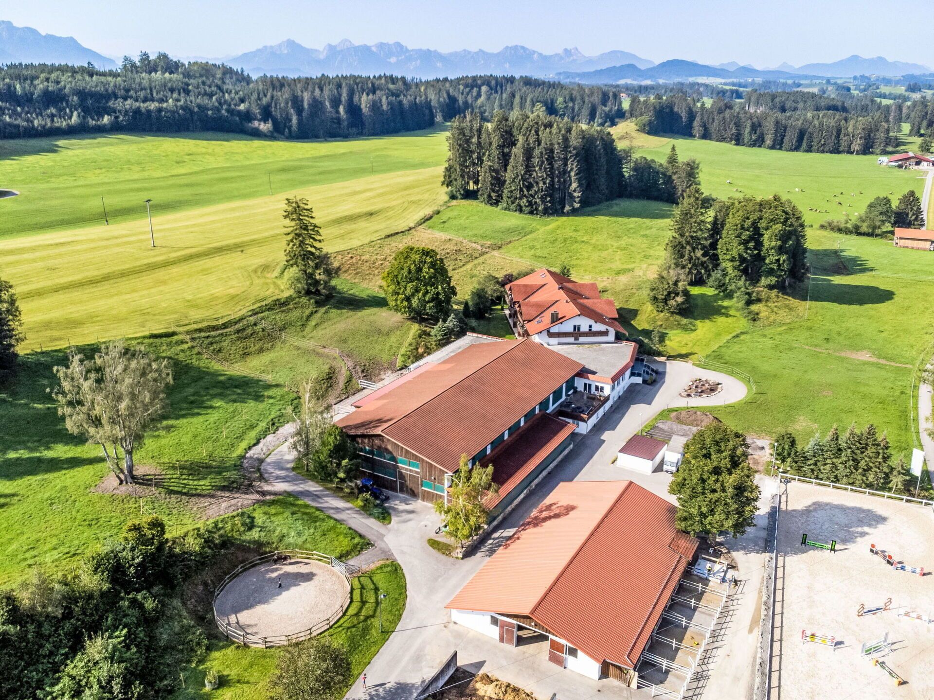 Eine Vogelperspektive eines Gebäudes mit rotem Dach, umgeben von grünen Feldern und Bäumen, mit Bergen im Hintergrund.