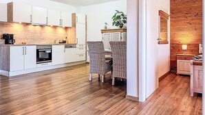 Una cocina moderna con armarios blancos, horno empotrado y encimera de madera. En la esquina, una silla de mimbre y una planta en maceta.