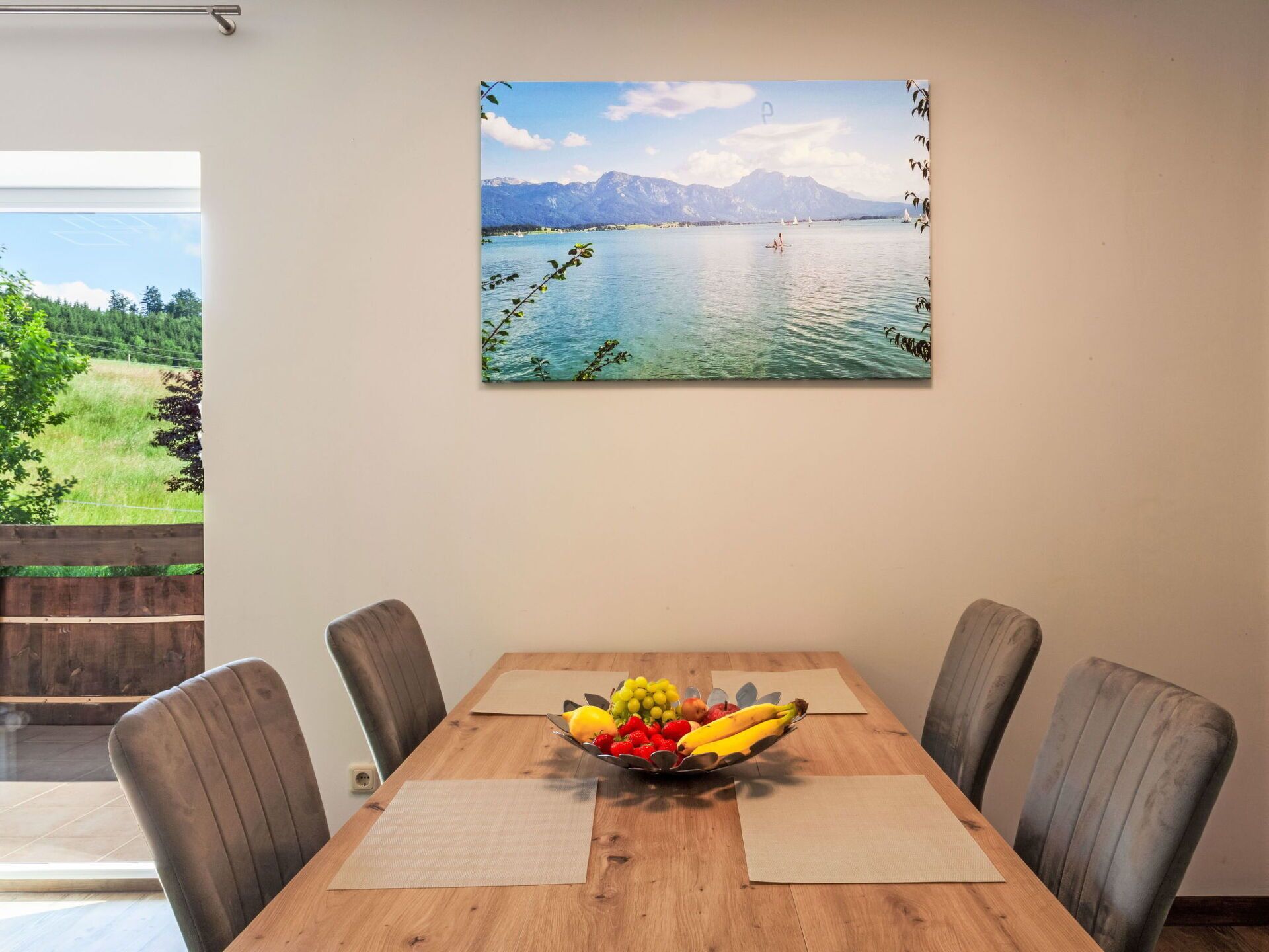 A dining table with a fruit bowl and placemats, a painting of a lake and mountains, and a view of a green landscape through a window.