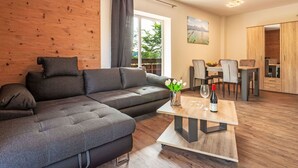 A modern living room with a sectional sofa, a wooden accent wall, a dining area with a table and chairs, and a view of a lake through the window. - Beautiful apartment for 4 guests with WIFI, TV, balcony, pets allowed and panoramic view (Klausmen)