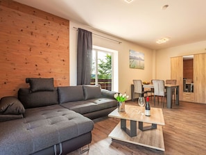Ein modernes Wohnzimmer mit einer Eckcouch, einer Wandpaneele aus Holz, einem Essbereich mit Tisch und Stühlen sowie Blick auf einen See durch das Fenster.