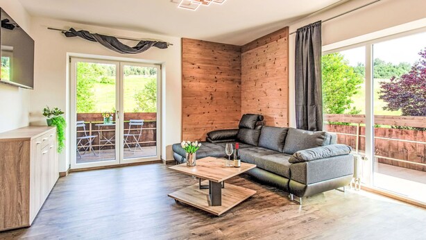 A modern living room with a grey sofa, wooden flooring, a coffee table, and a sliding glass door leading to an outdoor area with a wooden deck and chairs. - Beautiful apartment for 4 guests with WIFI, TV, balcony, pets allowed and panoramic view (Klausmen)
