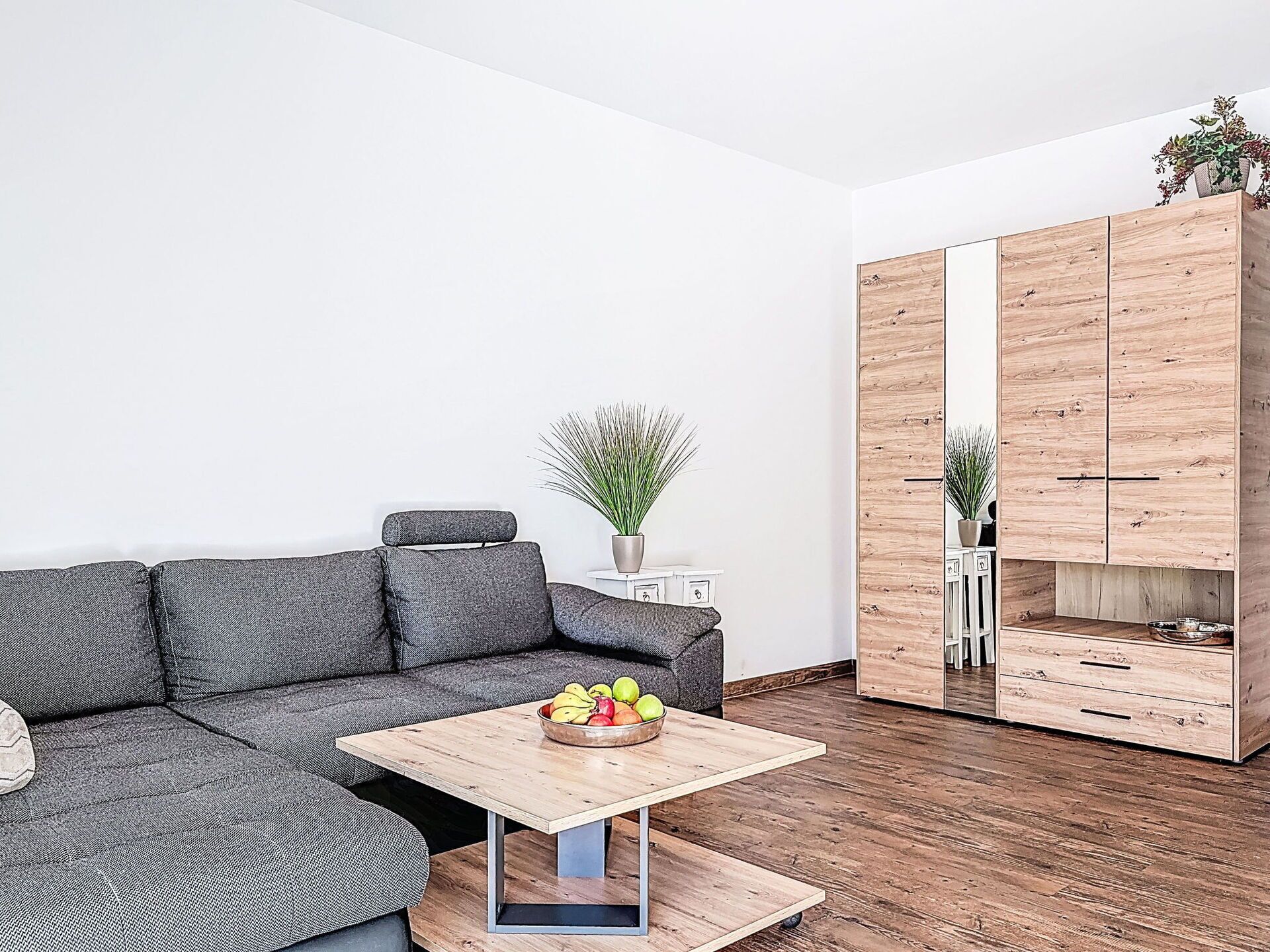 Una sala de estar moderna con un sofá seccional gris, una mesa de centro de madera con fruta, una mesita de noche blanca y un armario de madera con espejo.