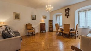 Un vaste salon doté d’un parquet en point de Hongrie, d’un lustre, d’un coin repas avec une table et des chaises, et d’une grande horloge murale.