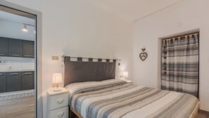 Une chambre à coucher comprenant un lit, des tables de chevet, un miroir, une fenêtre avec des rideaux et une vue sur la cuisine.