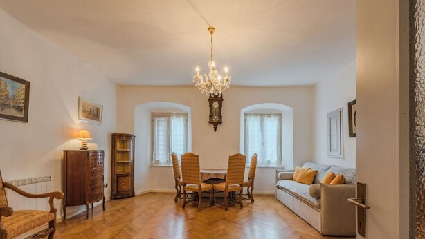 Un vaste salon doté d’un parquet en bois, d’un lustre, d’une table à manger avec des chaises, d’un canapé et d’une horloge ancienne accrochée au mur.