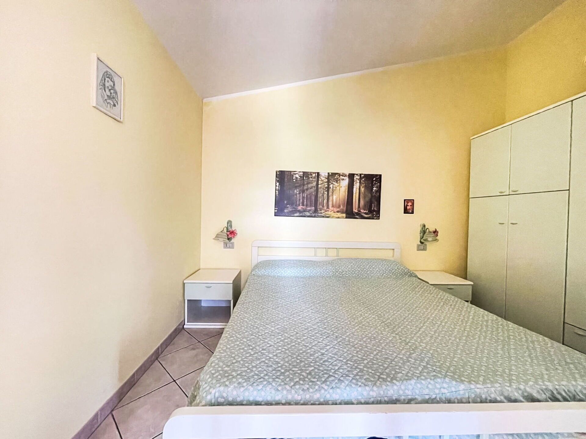 A bedroom with a bed, a nightstand, a wardrobe, and a framed picture on the wall.