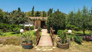 A pathway leading to a building with a pergola entrance, surrounded by a well-maintained garden with various plants and a clear blue sky. - Beautiful private villa for 5 people with A/C, TV, pets allowed and panoramic view (Villasimius)