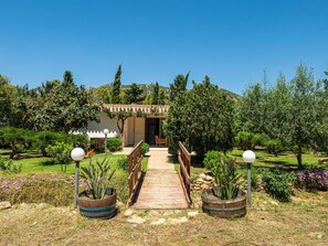 Ein Weg führt zu einem Gebäude mit Pergola-Eingang, umgeben von einem gepflegten Garten mit verschiedenen Pflanzen und blauem Himmel.
