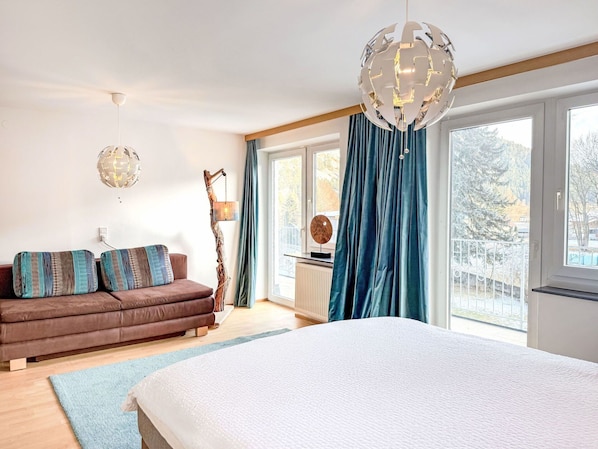 A bedroom with a large bed, a brown sofa with patterned cushions, a hanging light fixture, and a view of the outdoors through a window with blue curtains.