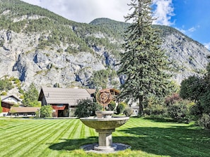 A mountain landscape with a stone fountain, a red-roofed building, and a large tree.