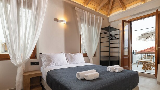 Une chambre à coucher avec un lit, des rideaux blancs, une table de chevet en bois et un balcon agrémenté de meubles en osier.