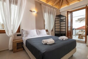 Een slaapkamer met een bed, witte gordijnen, een houten nachtkastje en een balkon met rieten meubilair.