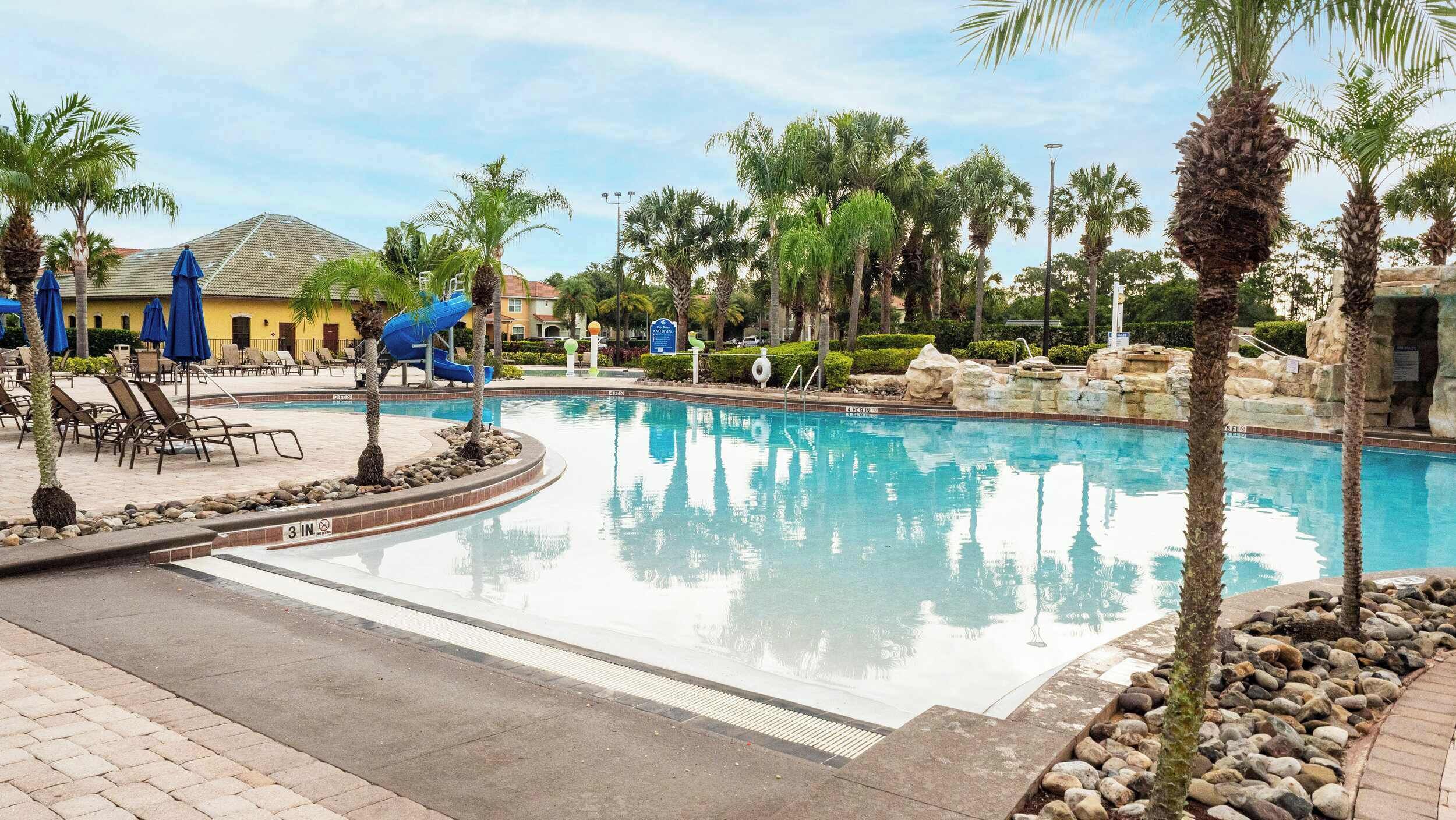 A swimming pool with lounge chairs and umbrellas, surrounded by palm trees and a building in the background.