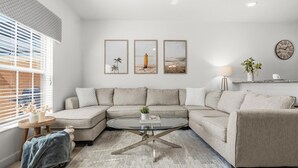 Una sala de estar moderna con un sofá modular, una mesa de centro de vidrio y tres cuadros con motivos playeros en la pared.