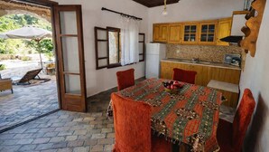 A dining area with a table covered by a patterned tablecloth, wooden chairs, and a view of the outdoors through an open door. - Saltos Apartment 2 - Pool Garden Getaway (Nea Propontida)