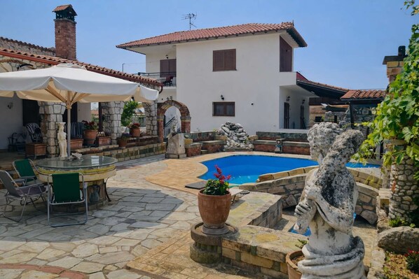 A villa with a swimming pool, stone patio, outdoor furniture, and a statue. - Saltos Apartment 2 - Pool Garden Getaway (Nea Propontida)