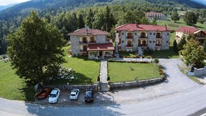 A stone building with a red-tiled roof, surrounded by a well-maintained lawn and parked cars. - Orama Hotel - Lake Plastira Fireplace Suite (Lake Plastiras)