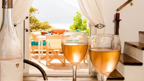 A table with a bottle and two glasses of wine, a painting of a person on a beach, and a view of a mountainous landscape through a window with curtains. - Casa Azul Villa - Seafront Getaway in Kalymnos (Kalymnos)