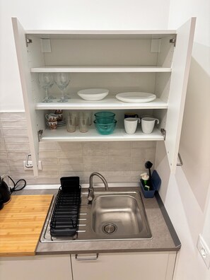 A kitchen with a built-in sink, a stove, and shelves above holding various dishes and glassware. - Apartment Monza Station I 15 min Milan City Center (Monza)