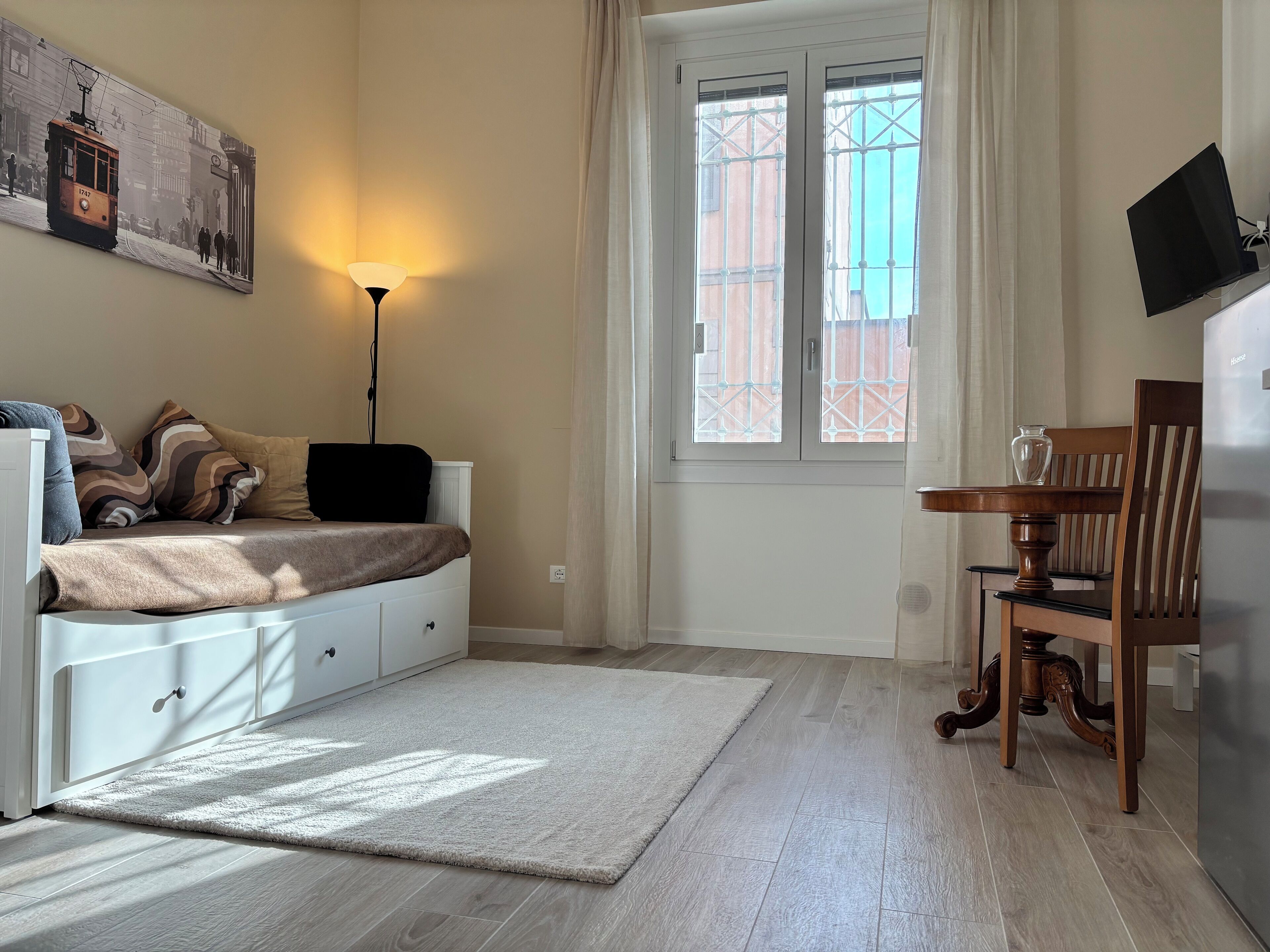 A bedroom with a bed, a desk, a chair, a lamp, and a window with a view of buildings.