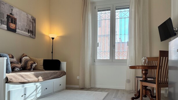 Ein Schlafzimmer mit Bett, Schreibtisch, Stuhl, Lampe und einem Fenster mit Blick auf Gebäude.