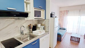 Een compacte keuken met een spoelbak, kookplaat, magnetron en koelkast. Er is een klein eethoekje met een tafel en stoelen.