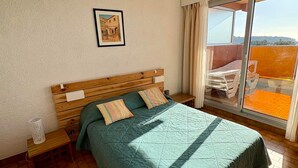Ein Schlafzimmer mit einem hölzernen Bett, einem kleinen Nachttisch, einem Fenster mit Vorhängen und einem Balkon mit Tisch und Stühlen.