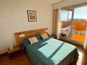 A bedroom with a wooden bed, a small bedside table, a nightstand, a window with curtains, and a balcony with a table and chairs. - Apartment for 8, spacious living with Wi-Fi included (Roquebrune-Cap-Martin)