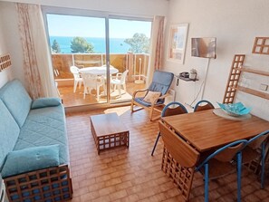 Ein Zimmer mit einer Couch, einem Esstisch mit Stühlen, einem Fernseher und einem Balkon mit Meerblick.
