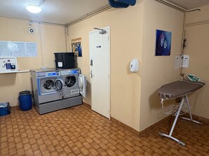 Une buanderie équipée d’un lave-linge et d’un sèche-linge à chargement frontal, d’une planche à repasser et d’une poubelle bleue.