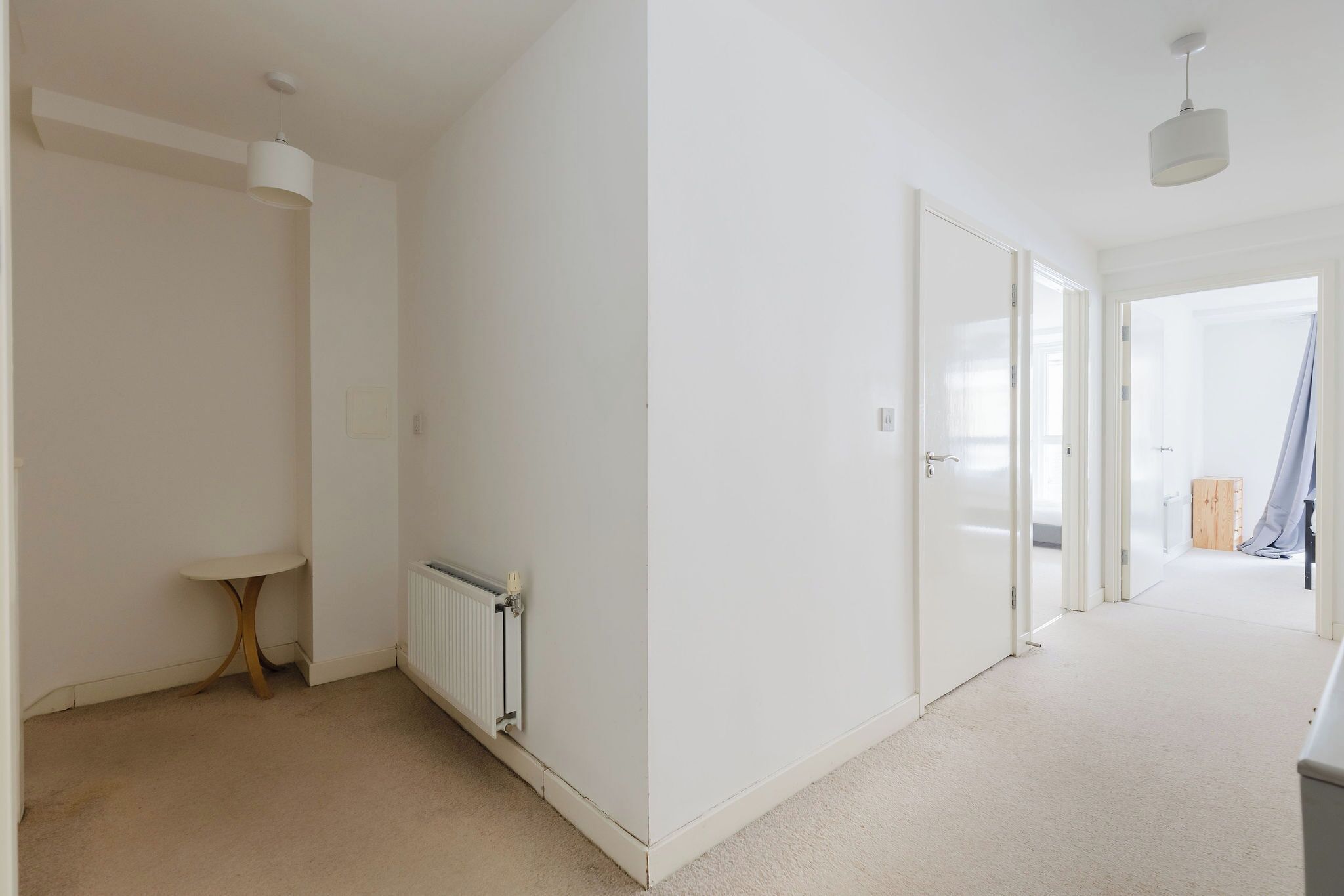 A hallway with white walls, a radiator, a small table, and a door leading to another room.