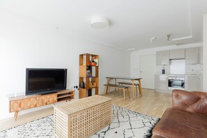 A modern living room with a flat-screen TV, wooden entertainment unit, a high shelf with books, a dining table with chairs, and a kitchen area with a stove and oven. - Calming 2BDR Hideaway 25 Mins From Tower Bridge (Greater London)