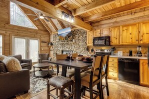 A cozy cabin interior with a stone wall, wooden ceiling, a dining area, and a kitchen with wooden cabinets and appliances. - Close to Gatlinburg, 3 bedroom, 2 bath, hot tub, pool table (Gatlinburg)
