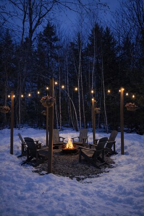 A snow-covered outdoor seating area with chairs and a fire pit, surrounded by trees and string lights. - Kid Friendly Cabin, Mount Snow 8 Min, Hot Tub, Game Room, Fire Pit, Dogs Ok (Dover)