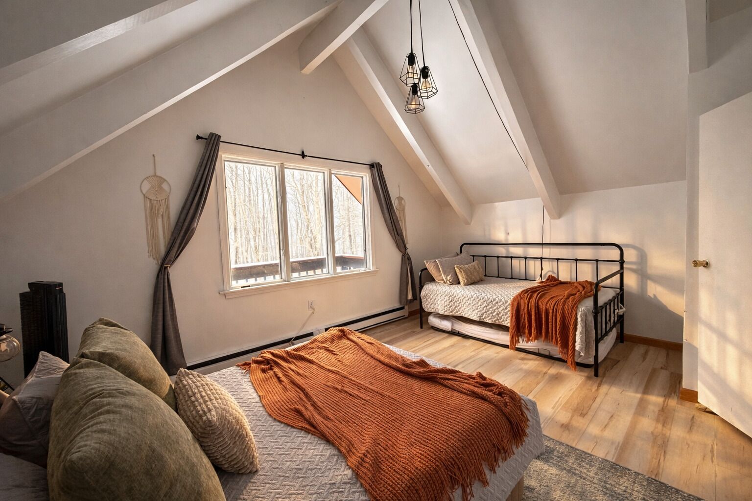Dormitorio tipo loft con techo inclinado, una ventana grande, una cama, un sofá y una lámpara colgante.