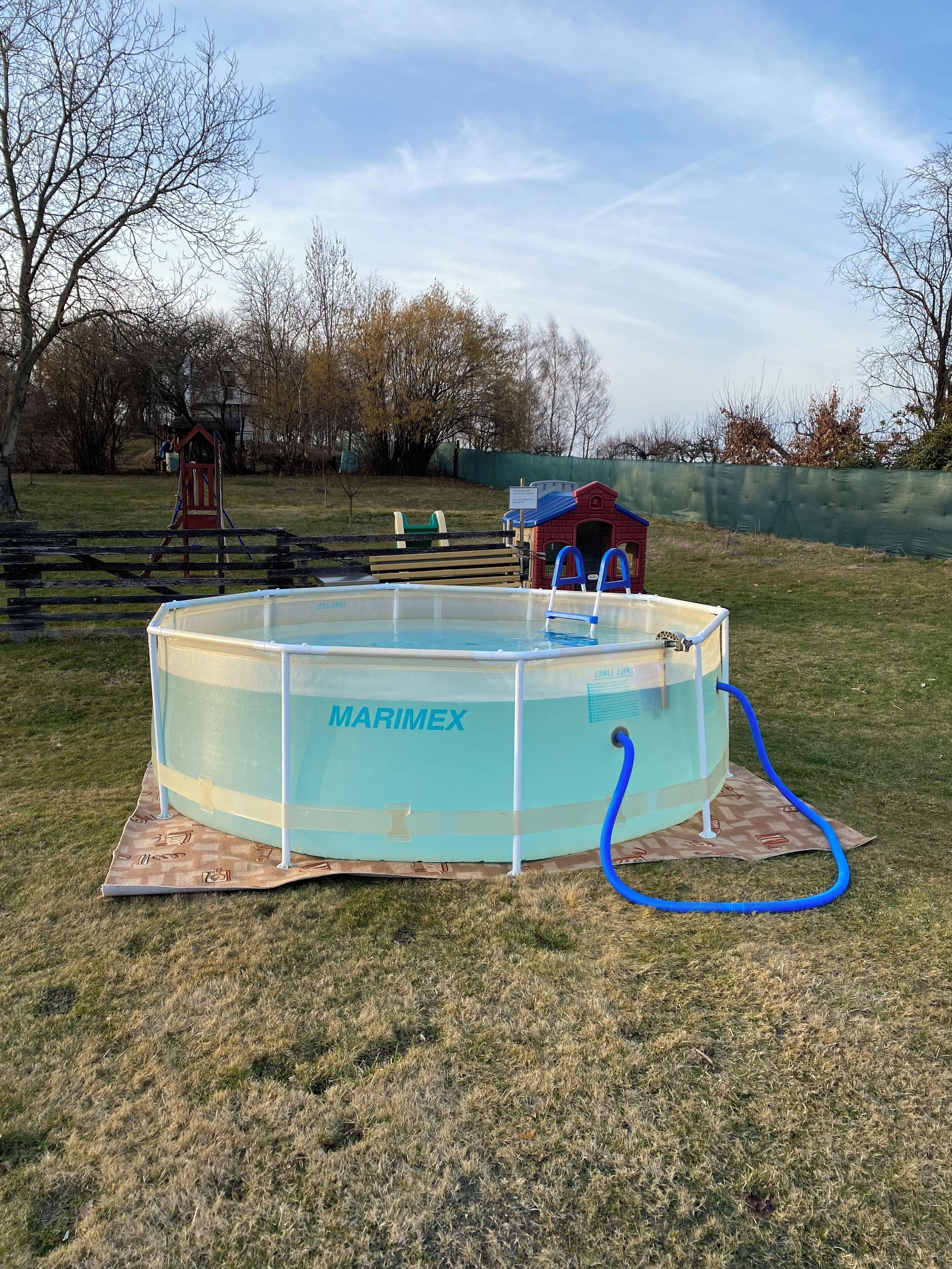 Una piscina rodona amb una mànega blava connectada, situada en una zona verda amb una terrassa de fusta a prop.