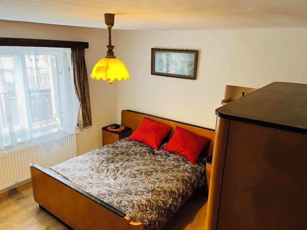 A bedroom with a bed, a nightstand, a lamp, a radiator, a window with curtains, and a framed picture on the wall.