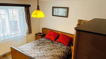 A bedroom with a bed, a nightstand, a lamp, a radiator, a window with curtains, and a framed picture on the wall.
