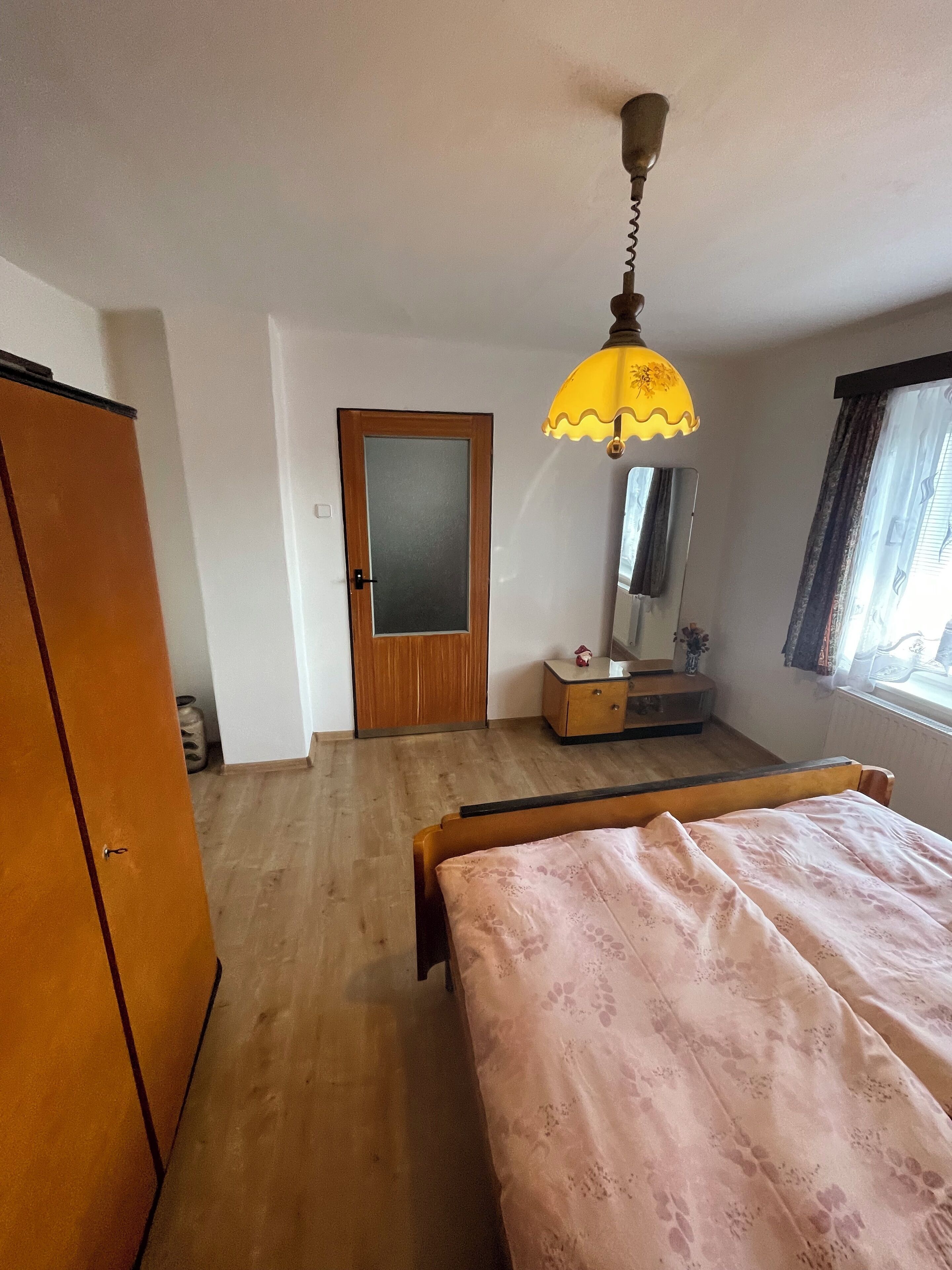 A bedroom with a wooden wardrobe, a bed with a floral bedspread, a small bedside table with a mirror, and a hanging lamp.