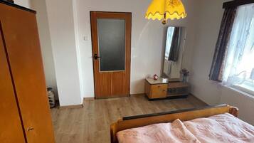 A bedroom with a wooden wardrobe, a bed with a floral bedspread, a small bedside table with a mirror, and a hanging lamp.