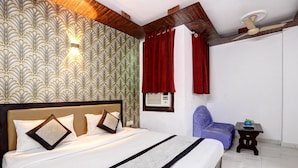 A hotel room with a large bed, a headboard, a wall-mounted light, a ceiling fan, a window with red curtains, a small table with a plant, and a blue chair. - Hotel Delhi Darbar-Near Karolbagh Metro (New Delhi)