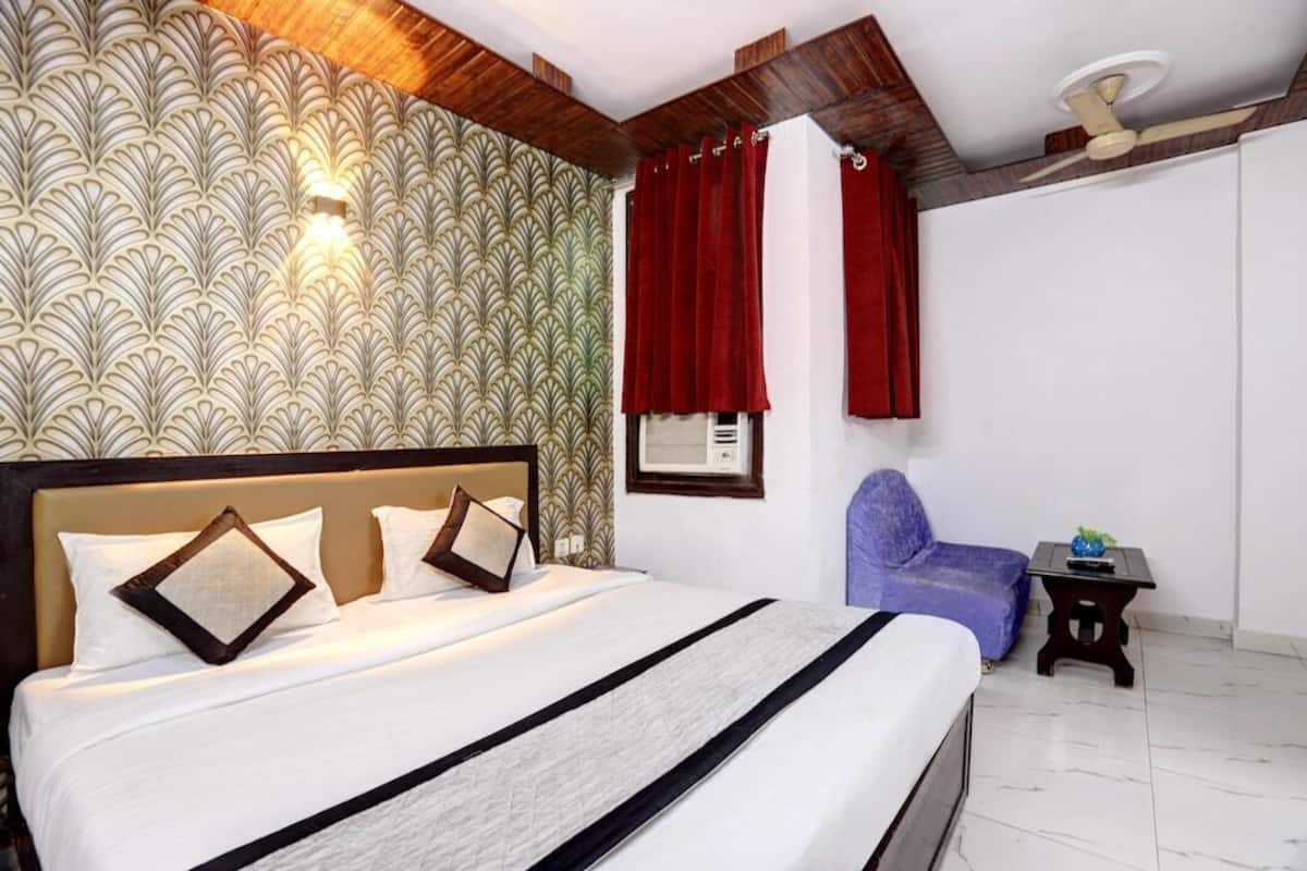Habitación de hotel con una cama grande, cabecera, lámpara de pared, ventilador de techo, ventana con cortinas rojas, una pequeña mesa con una planta y una silla azul.