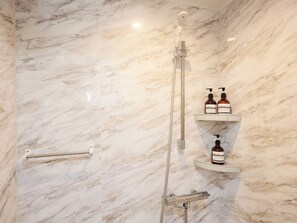 A bathroom with a marble wall, a shower with a handheld showerhead, and shelves with bottles of shampoo and conditioner.