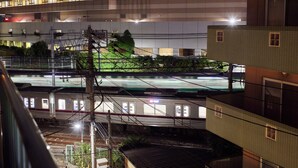 Un andén de estación de tren con un tren en movimiento, cables aéreos y un edificio con múltiples ventanas y balcones.