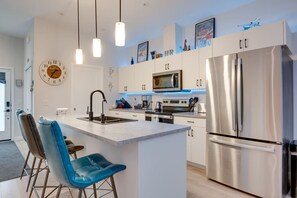 Uma cozinha moderna com ilha central, eletrodomésticos de aço inoxidável, luminárias pendentes e um relógio na parede.