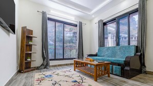 A living room with a sofa, a coffee table, a wooden shelf, and a large window with curtains. - Alaya Stays Vyasa Duplex With Views (Mussoorie)