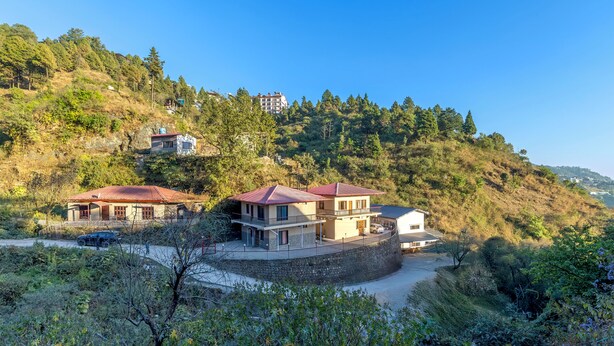 A hillside property with multiple houses, featuring a stone retaining wall, a paved driveway, and a mix of residential buildings. - Alaya Stays Vyasa Duplex With Views (Mussoorie)
