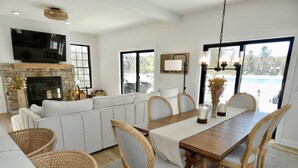 Uma sala de estar ampla com um sofá seccional grande, uma mesa de jantar com cadeiras, cestos de vime, uma lareira e uma televisão fixada na parede.